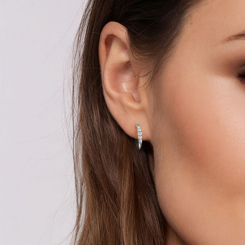 Close-up of a woman's ear wearing Gabriel & Co 14K White Gold Classic Round 15mm Diamond Huggie Earrings. Her brown hair is tucked behind her ear, and she has smooth, glowing skin with subtle makeup.