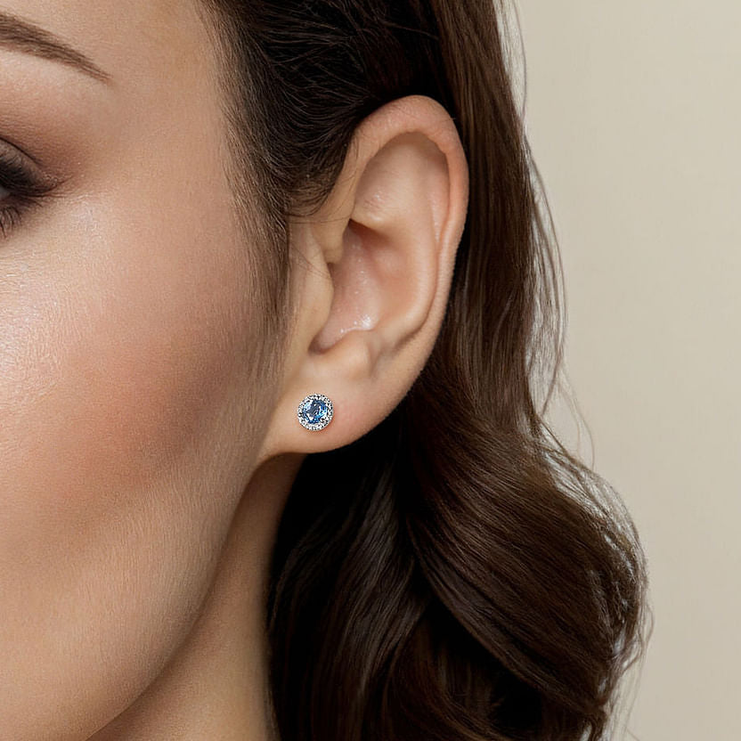Close-up of a woman's ear with wavy brown hair, wearing Gabriel & Co 14K White Gold Diamond Halo Swiss Blue Topaz Stud Earrings. The background is plain and light-colored.