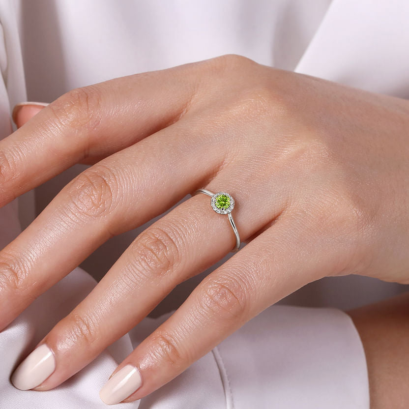A hand with manicured nails wears the Gabriel & Co 14K White Gold Peridot and Diamond Halo Promise Ring, showcasing a round green peridot and sparkling diamond halo, set against a light, silky fabric background.