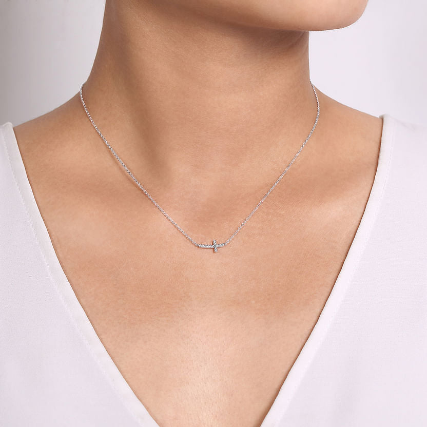 A person wears the Gabriel & Co 14K White Gold Sideways Curved Diamond Cross Necklace, featuring a small cross pendant, against light skin and a white V-neck top with a plain, neutral background.