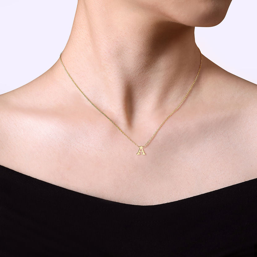 Wearing an off-the-shoulder black top, a person showcases the Gabriel & Co 14K Yellow Gold A Initial Necklace. The background is plain and light-colored.