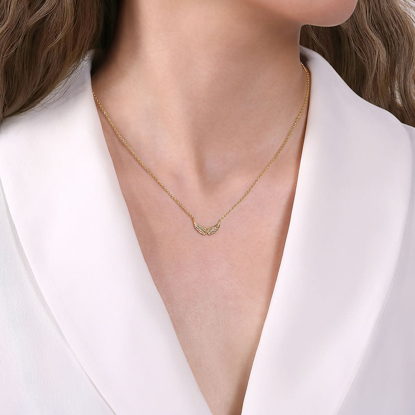 A woman wears a white blouse and showcases the Gabriel & Co 14K Yellow Gold Angel Wings Necklace, its delicate pendant elegantly resting below her collarbone, drawing attention to the necklace and her neckline.