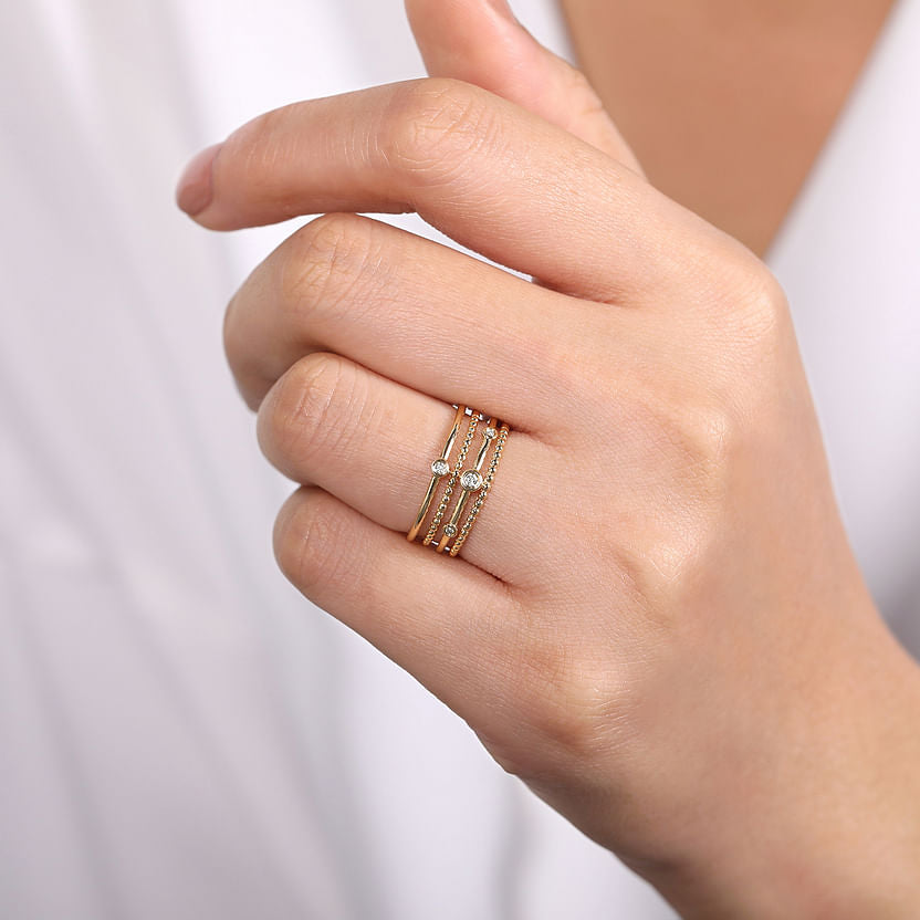 A hand with manicured nails showcases the Gabriel & Co 14K Yellow Gold Bezel Set Diamond Station Layered Ring, featuring three diamond-accented bands. The person is dressed in a white garment.