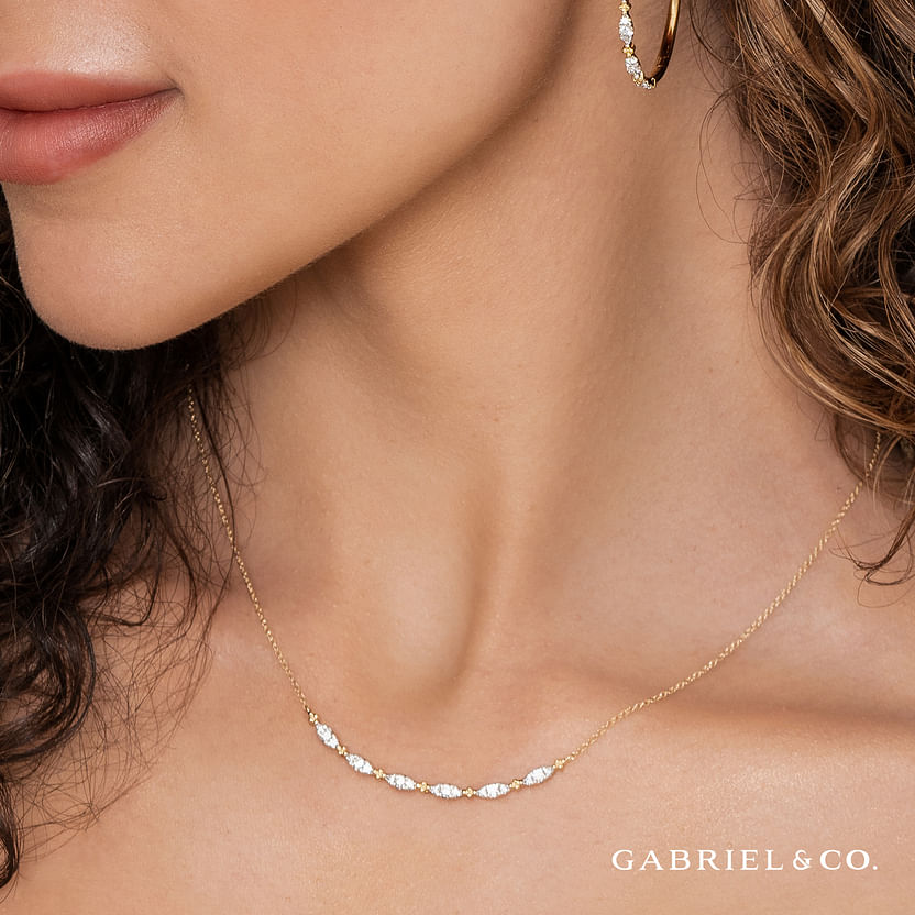 A woman wears the Gabriel & Co 14K Yellow Gold Curved Bujukan Bar Diamond Necklace and matching hoop earring. The image highlights her neck, jewelry, and lower face against a neutral background with "Gabriel & Co." in the corner.