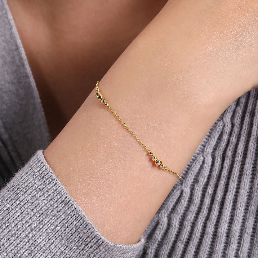 A close-up shows a wrist adorned with the Gabriel & Co 14K Yellow Gold Graduating Bead Stations Bracelet, elegantly paired with a light gray ribbed long-sleeve top.