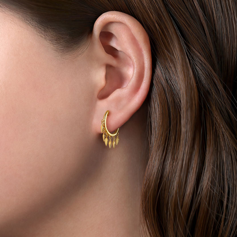 A close-up of a woman's ear wearing Gabriel & Co 14K Yellow Gold Huggie Earrings with spike drops and leaf-shaped dangles, set against brown hair.