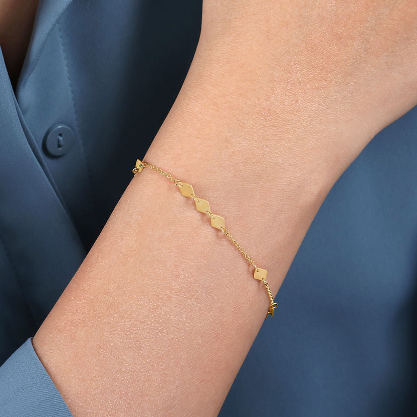 A person wearing the Gabriel & Co 14K Yellow Gold Rhombus Stations Bracelet with small, diamond-shaped charms on their wrist, styled with a blue long-sleeve shirt—a perfect geometric jewelry accent.