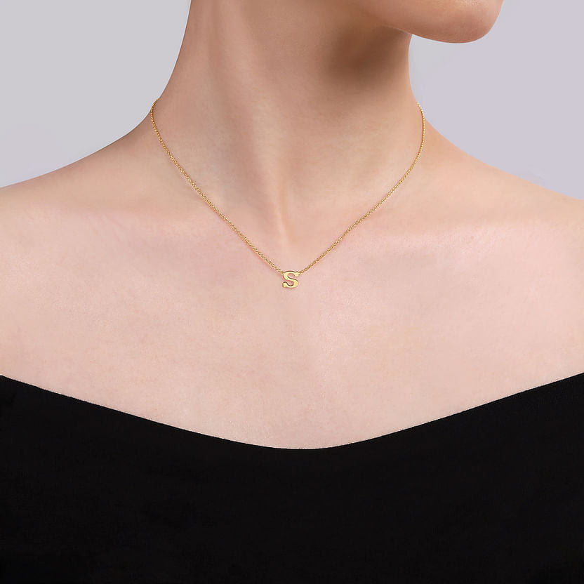 A person wearing an off-the-shoulder black top and a Gabriel & Co 14K Yellow Gold S Initial Necklace, featuring a pendant shaped like the letter "S.