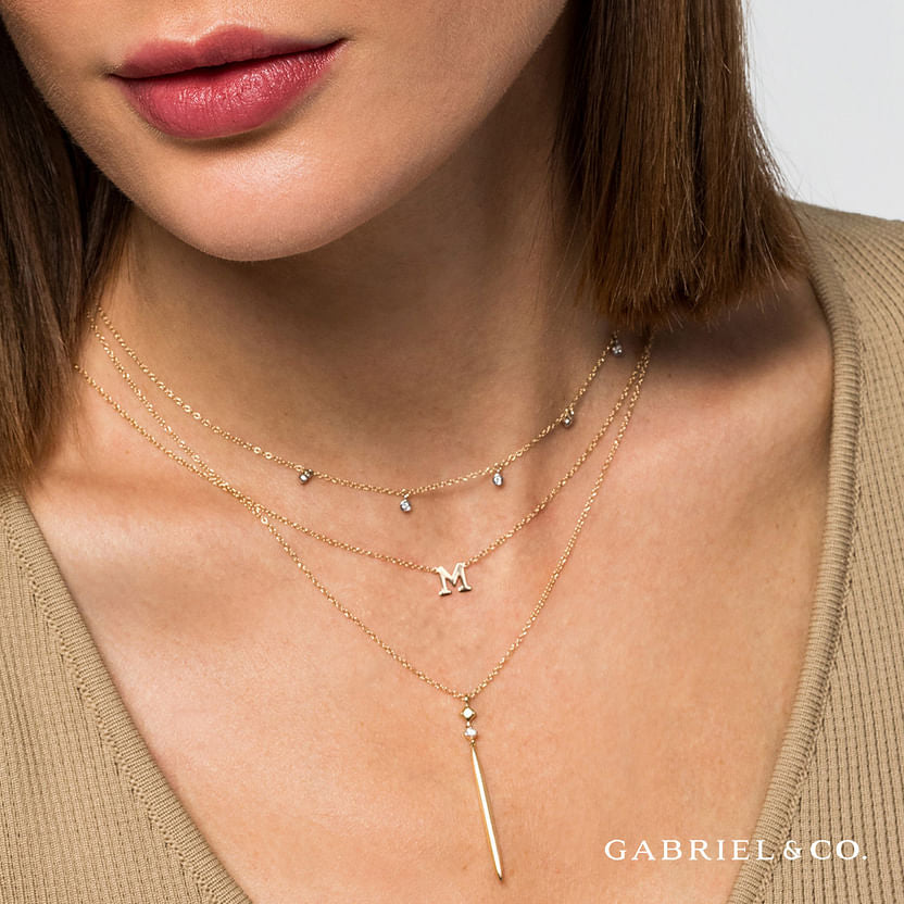 A woman with light skin and straight brown hair wears the 14K Yellow-White Gold Diamond Stations Droplet Necklace from Gabriel & Co, layered with two other necklaces. She has a beige top. "Gabriel & Co." text appears in the corner.
