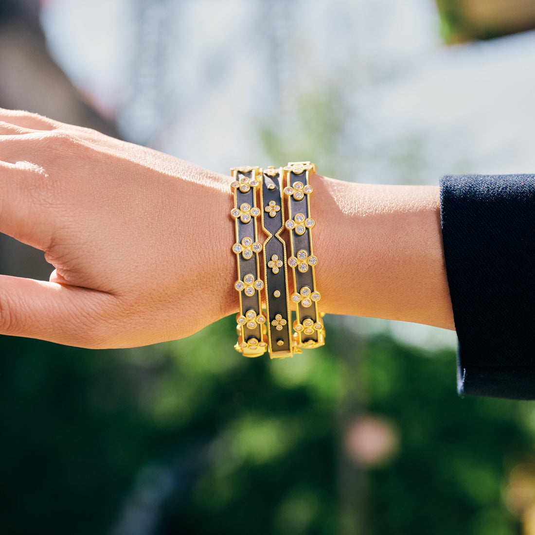 A hand wears Freida Rothman's Signature Brooklyn in Bloom Clover Hinge Bangles, gold adorned with intricate floral designs and gemstones, against blurred greenery. The sleeve reflects Brooklyn's spirit in bloom.