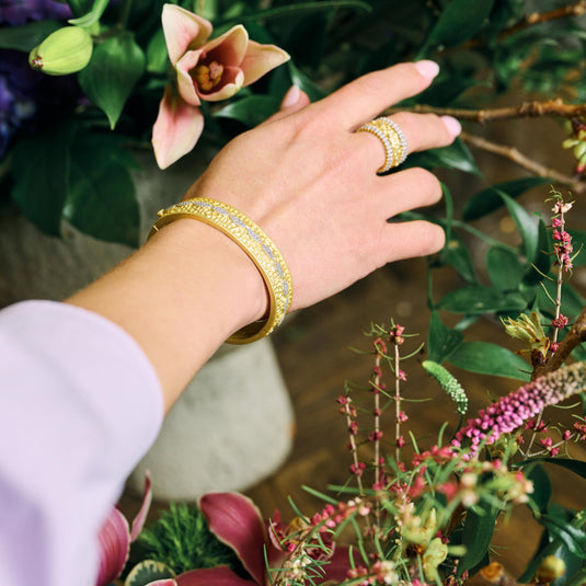 A hand adorned with the Freida Rothman Armor of Hope City Blooms Hinge Bangle and ring reaches for vibrant flowers. The 14K matte hammered gold jewelry, ornate with sparkling details, perfectly complements the colorful floral display.