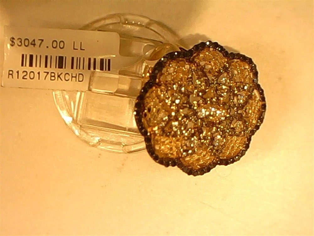 A Ryan Gems 14k yellow gold large flower ring with black, champagne, and white diamonds (SI) sits on a clear stand. Attached is a "$3047.00" price tag labeled "R12017BKCHD.
