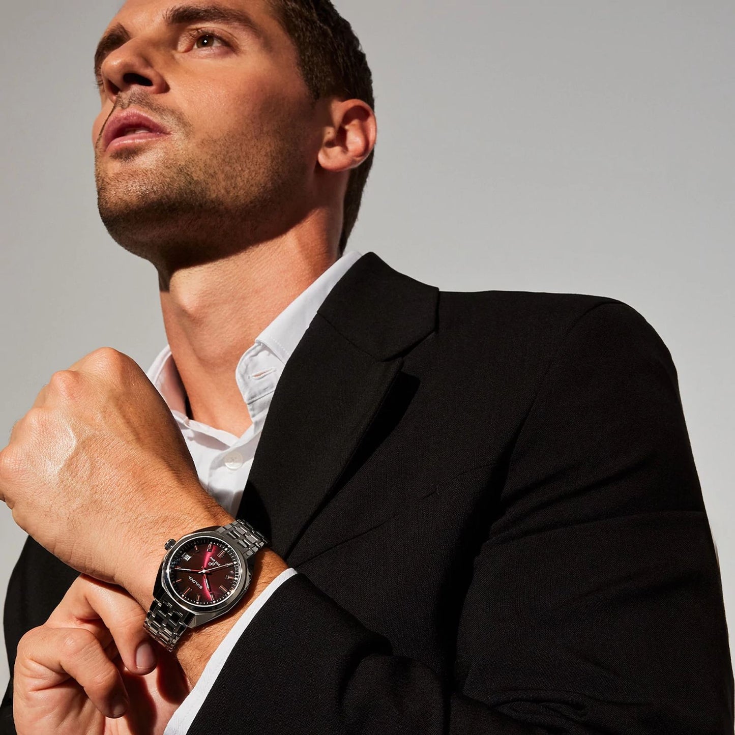 A man in a black suit and white shirt looks upward, showcasing the Bulova Watch Company’s Jet Star Watch with Maroon Dial and Stainless Steel Bracelet on his left wrist against a well-lit plain background.