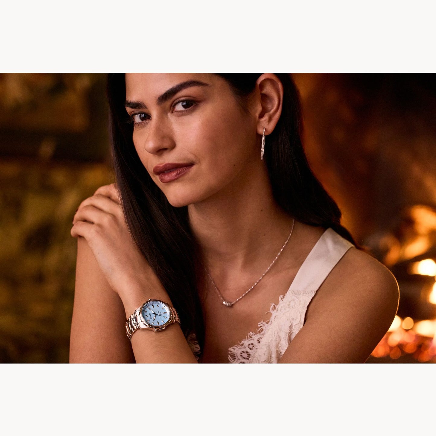 A woman with long dark hair wears a white lace top, hoop earrings, and a Bulova Watch Company Marine Star Ladies' Watch with mother of pearl and diamonds. She stands indoors with her arms crossed in warm lighting.