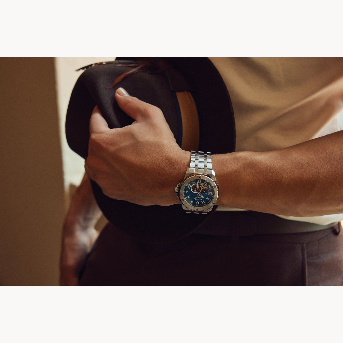 A person dressed in a white shirt and brown trousers is shown holding a dark-colored hat in one hand. They are wearing a Bulova Watch Company Marc Anthony Blue Dial Automatic Watch, featuring diamond hour markers and a silver wristwatch with a blue face on the same hand. The background is softly lit and out of focus.