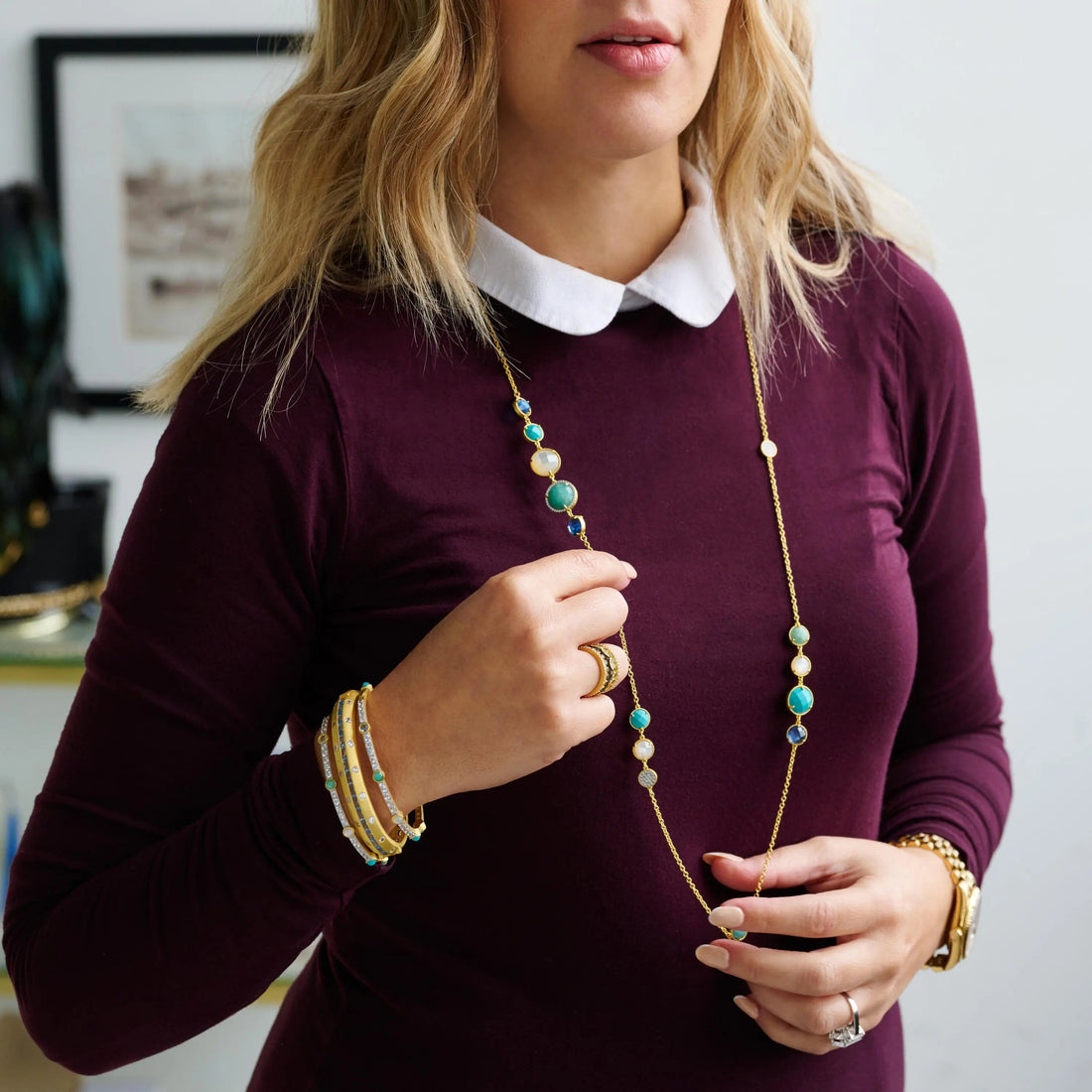 In a blurred room, a person in a maroon dress with a white collar showcases the Freida Rothman Shades of Hope Long Chain Necklace, featuring turquoise and white beads. They accessorize with several bracelets and a watch on their left wrist, with an indistinct picture frame in the background.