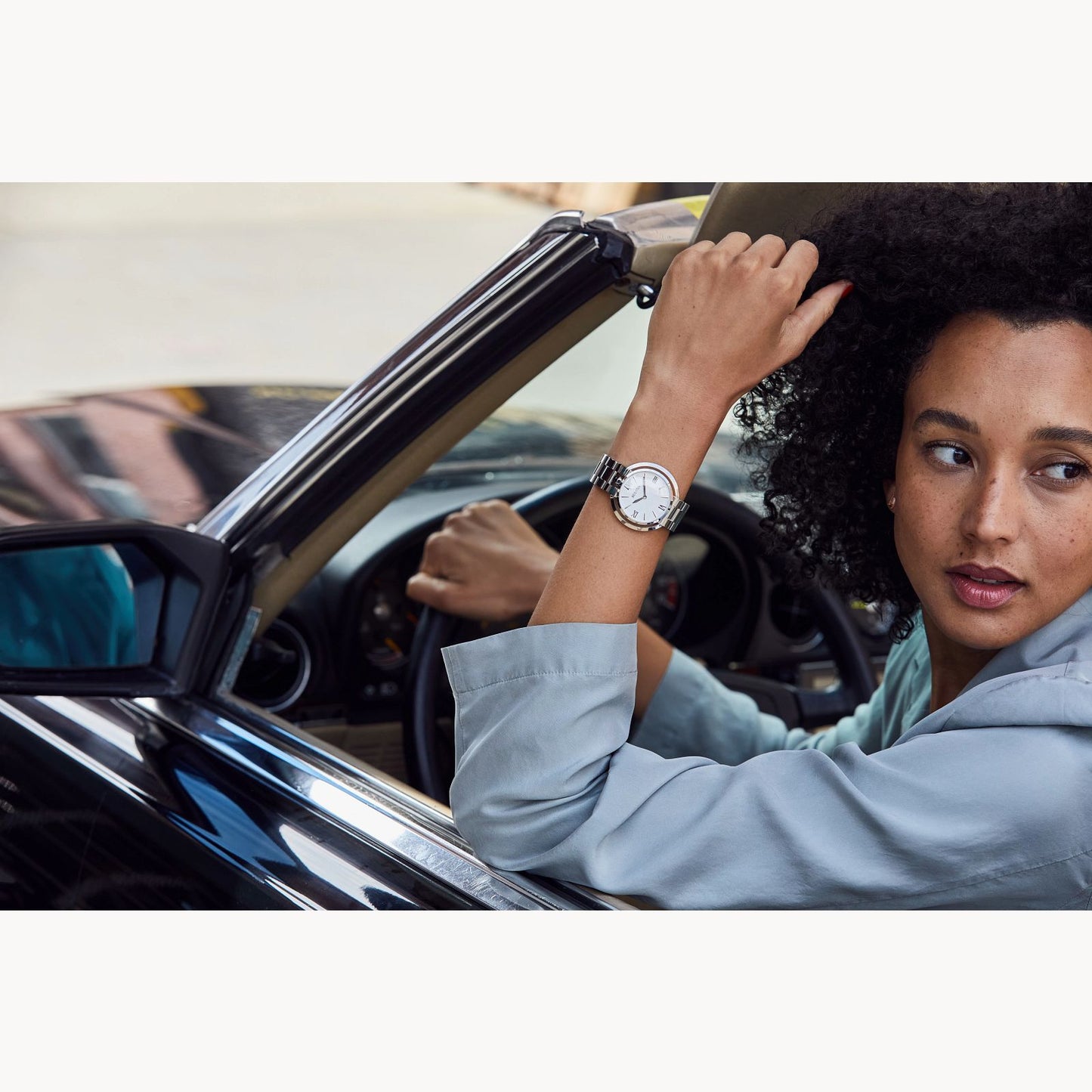 A woman with curly hair, wearing a light blue top and the Bulova Rubaiyat Women's White Dial Stainless Steel Watch by Bulova Watch Company, sits in a convertible, looking over her shoulder with her hand resting on the car’s frame.