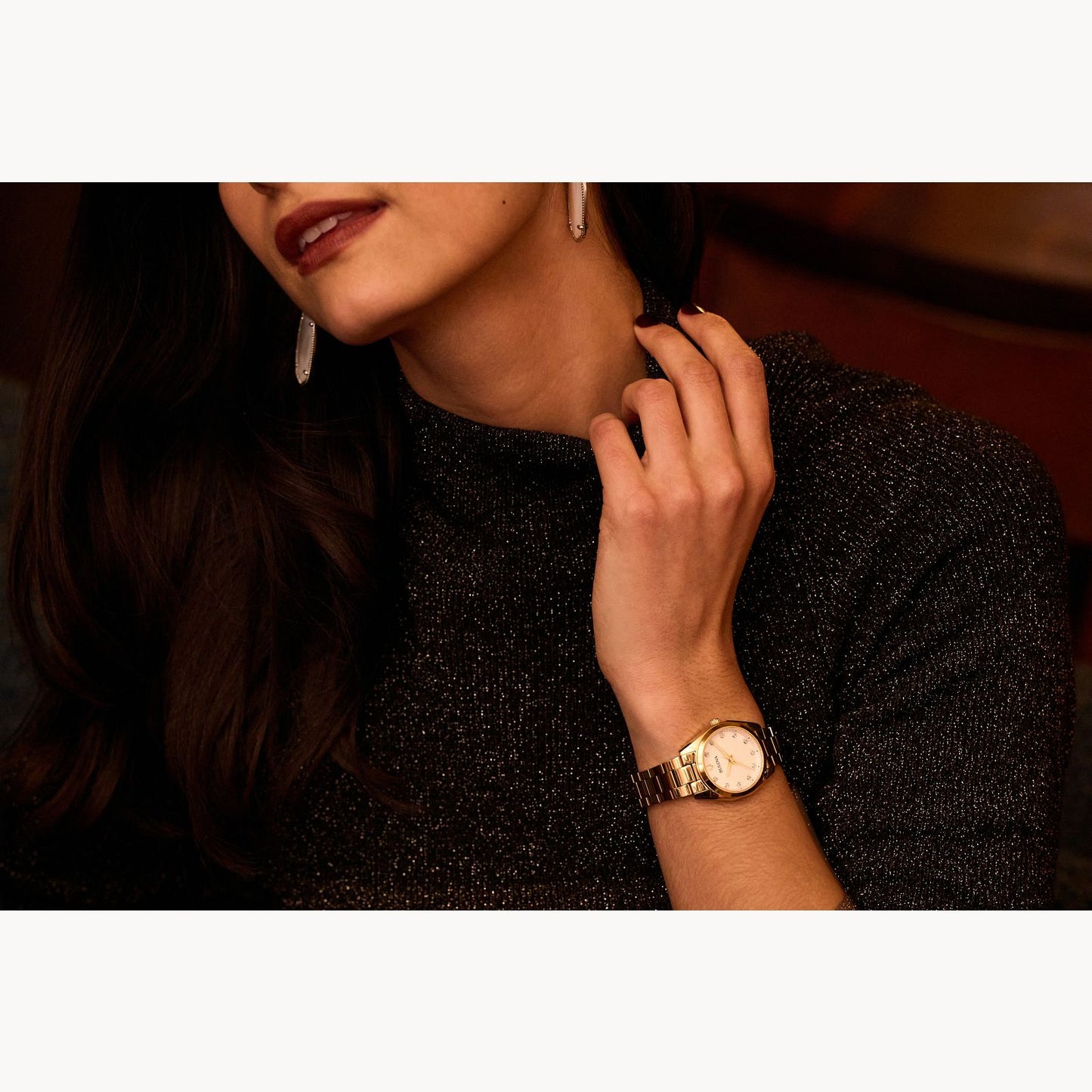 A woman wearing a dark, sparkly top and drop earrings touches her neck, showcasing the Bulova Watch Company Ladies Surveyor in stainless steel with a sapphire crystal and MOP dial. Her maroon lipstick and dark hair are partially visible.