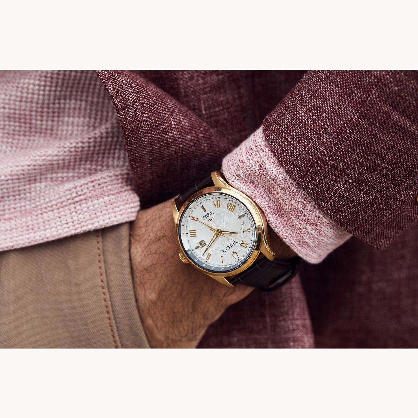 A person wears a Bulova Wilton Automatic Watch with leather strap by Bulova Watch Company, featuring a white dial, Roman numerals, and stainless steel case, paired with a pink textured shirt, burgundy jacket, and light brown pants.