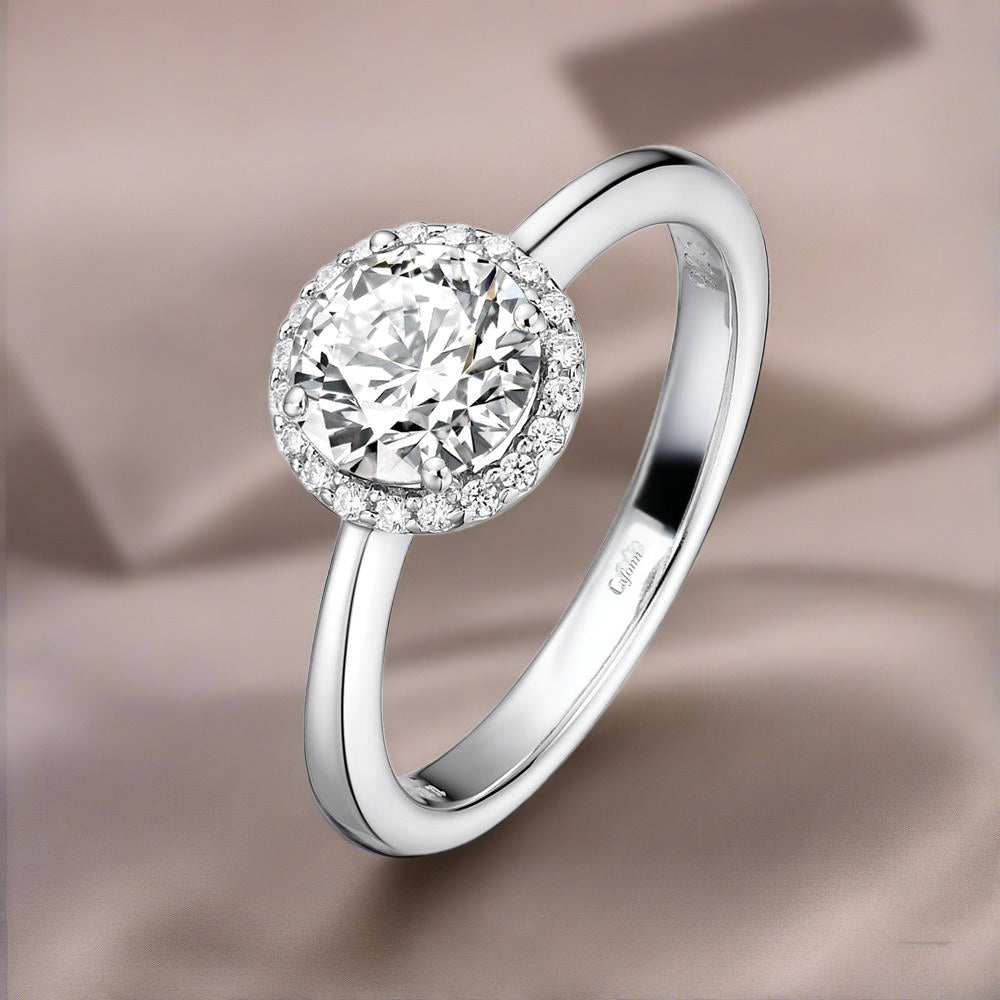 An elegant Lafonn April Birthstone Ring, featuring a round-cut simulated diamond haloed by smaller diamonds, set on a sleek platinum band. The soft, blurred beige fabric background enhances the ring's brilliance.