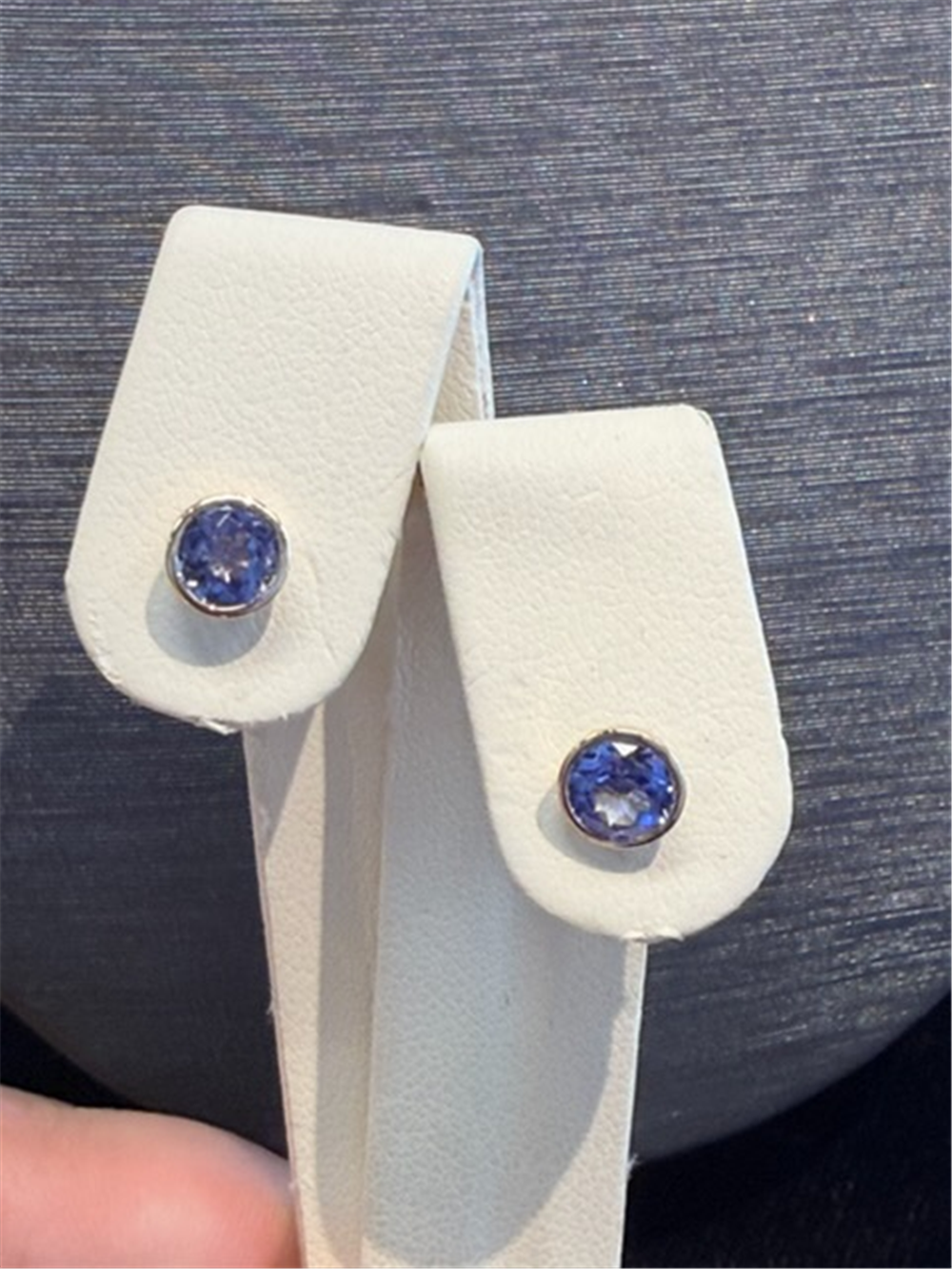 A pair of Tanzanite bezel set studs in 14k yellow gold by Ryan Gems, displayed on a white leather holder against a gray background.