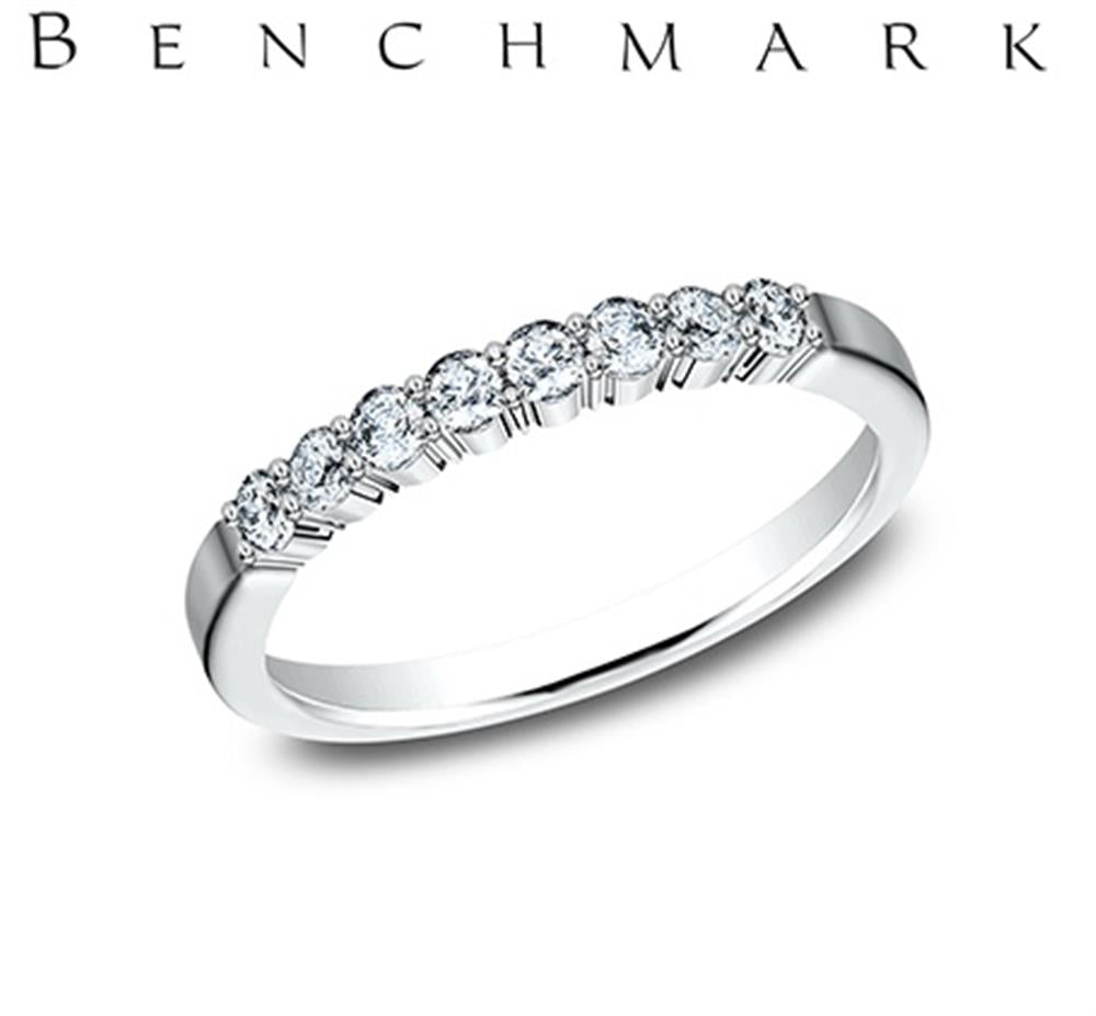 The 14K White Gold 0.32cts Lab Grown Diamond Shared Prong Set Band by Benchmark Rings features a row of small, round lab-grown diamonds set along the top half. The brand name "Benchmark Rings" is elegantly displayed in black lettering at the top of the image, enhancing its simple and elegant design.