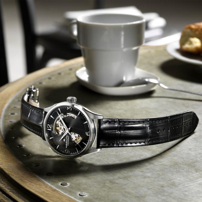 A Hamilton Jazzmaster Automatic Open Heart Watch with a black leather strap sits on a table next to a white coffee cup and saucer. The watch face, showcasing intricate mechanical details powered by Swiss automatic movement, is set against a background featuring a blurred chair and pastry on a plate.