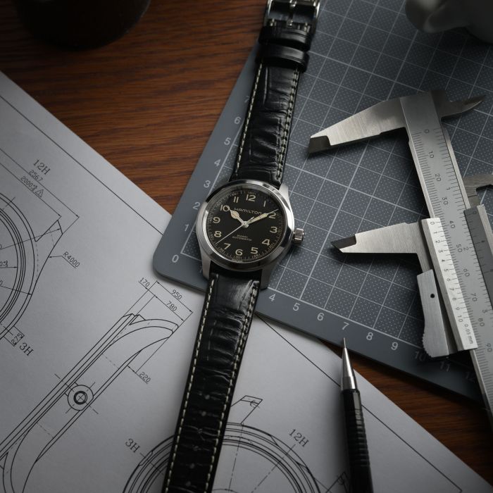 A black leather-strap wristwatch with an analog face, the Khaki Field Murph 38mm by Hamilton, is placed on top of technical design sketches. Next to it are a pair of calipers, a pencil, and a cutting mat on a wooden desk, suggesting precision and craftsmanship in watchmaking.