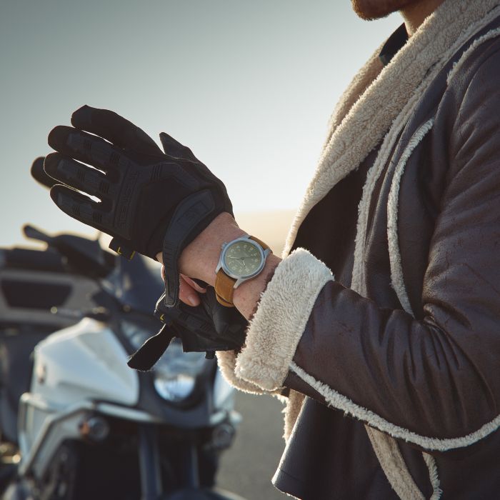 A person wearing a brown leather and shearling jacket puts on black gloves. They are standing near a parked motorcycle, with the focus on their wrists showing a Hamilton Khaki Field Titanium Automatic 42mm watch and their gloved hands. The scene is outdoors in daylight.