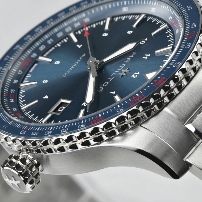 Close-up image of a Khaki Aviation Automatic Watch by Hamilton, showcasing its stainless steel wristband and striking blue dial. This automatic timepiece features a date display and 24-hour numerals, complemented by additional numbering on the bezel. The embossed star design on the crown adds a touch of elegance, while "Hamilton" and "KHAKI AVIATION CONVERTER" are prominently displayed on the dial.