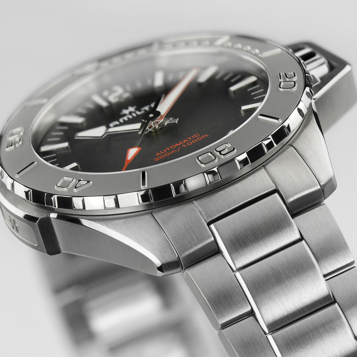 Close-up of the Khaki Navy Frogman Automatic 46mm by Hamilton, featuring a silver stainless steel band. The black watch face is adorned with white and orange hands and markers, displaying "Automatic 300m/1000ft" text. Ideal for maritime use, this timepiece has its bezel engraved with minute markers and is set to 3:35.