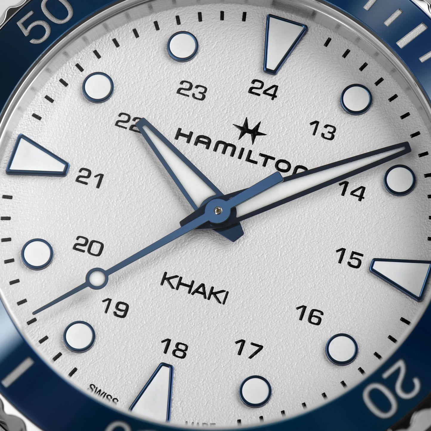 Close-up of the Hamilton Khaki Navy Scuba Quartz 37mm watch face showing a white dial with black numerals and blue hour, minute, and second hands. The outer ring features a striking blue bezel with white markers, and the watch displays both 12-hour and 24-hour formats.