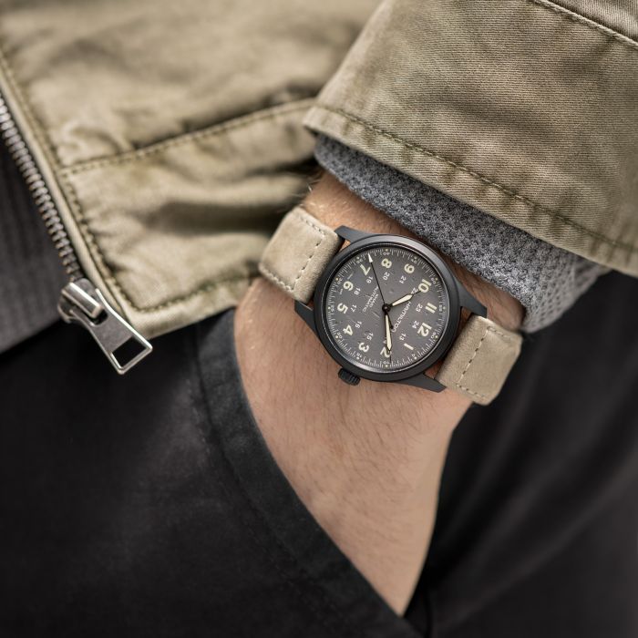 A close-up of a person's wrist wearing the Hamilton Khaki Field Titanium Automatic 38mm watch, which features a tan fabric strap and white numbering on the face. The person is dressed in a grey sweater under a beige jacket with a metal zipper, and their hand is placed in the pocket of black pants.
