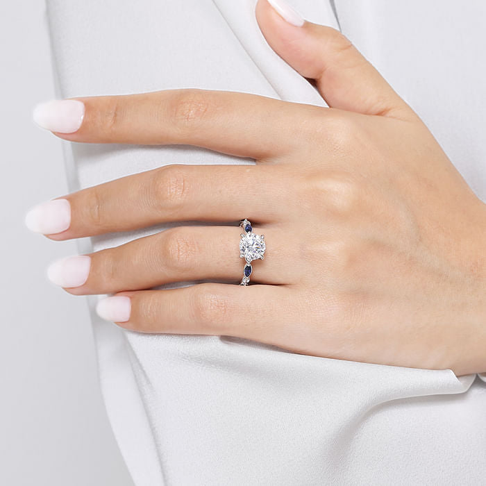 A manicured hand rests on white fabric, showcasing Gabriel & Co’s 14K White Gold Round Sapphire and Diamond Engagement Ring | 2 carat—an elegant design with a central gemstone and two blue sapphires.