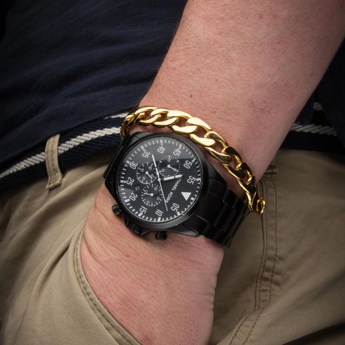 A person is wearing an INOX Stainless Steel Gold IP Diamond Cut Curb Chain Bracelet and a sleek black watch with a large dial. Their hand rests in the pocket of beige pants, and they are dressed in a dark shirt with a white and navy striped belt visible.