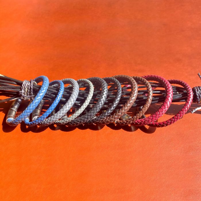 A set of nine braided leather bracelets, featuring the elegant 8mm Burgundy Genuine Leather Bracelet by INOX, arranged in a row on an orange background. The bracelets are available in a variety of colors including blue, brown, gray, black, and red. The texture of the leather and intricate braiding details are clearly visible.