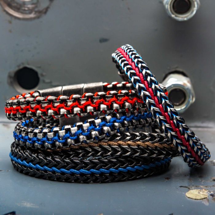 The Allegiance Red Stainless Steel Bracelet by INOX is stacked alongside other woven bracelets in an array of colors, including blue, black, and beige, against a metallic background adorned with bolts. These bracelets feature intricate patterns and metallic clasps, blending rugged elegance with a design reminiscent of antique brushed bold box links.