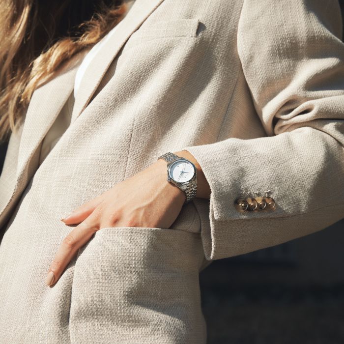 A person wearing a beige blazer with their left hand in the pocket. On their wrist is a Hamilton Lady Quartz Watch, featuring a white face and exuding elegance. The person's long hair and minimal fingers are visible against the fabric of the blazer, which is adorned with gold buttons on the sleeve for added sophistication.