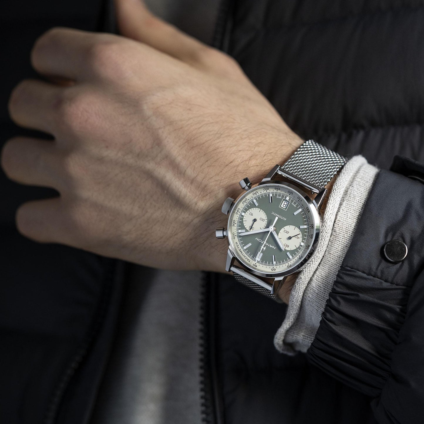 A person's wrist wearing the Hamilton Intra-Matic Automatic Chronograph 40mm watch, featuring a sleek, silver mesh band and a sophisticated grey dial with two subdials that evoke vintage charm. Their hand is partially visible, clad in a dark, puffy jacket with an inner sleeve in white and grey. The dark background highlights the elegance of the watch.