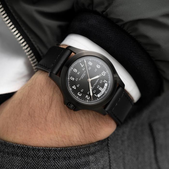 Close-up of a person wearing a Hamilton Khaki Field King Automatic Watch on their left wrist. The watch, steeped in military heritage, features a black round face with white numerals and a date display. The strap is black leather. The person is dressed in a grey herringbone-patterned garment, a white shirt, and a dark green jacket.