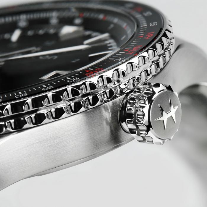 Close-up of a Hamilton Khaki Aviation Converter Automatic Watch, highlighting the stainless steel crown. The crown features a star emblem and is intricately detailed with fine grooves. The black dial with red and white markings is partially visible in the blurred background.