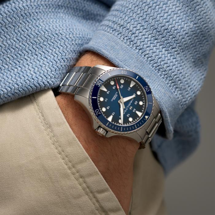 Close-up of a person's wrist wearing a Hamilton Khaki Navy Scuba Automatic Watch with a blue bezel and dial. The 43mm case adds to its sleek and modern design. Dressed in a light blue textured sweater and beige pants, their hand is tucked into the pocket. This watch boasts water resistance up to 300m.