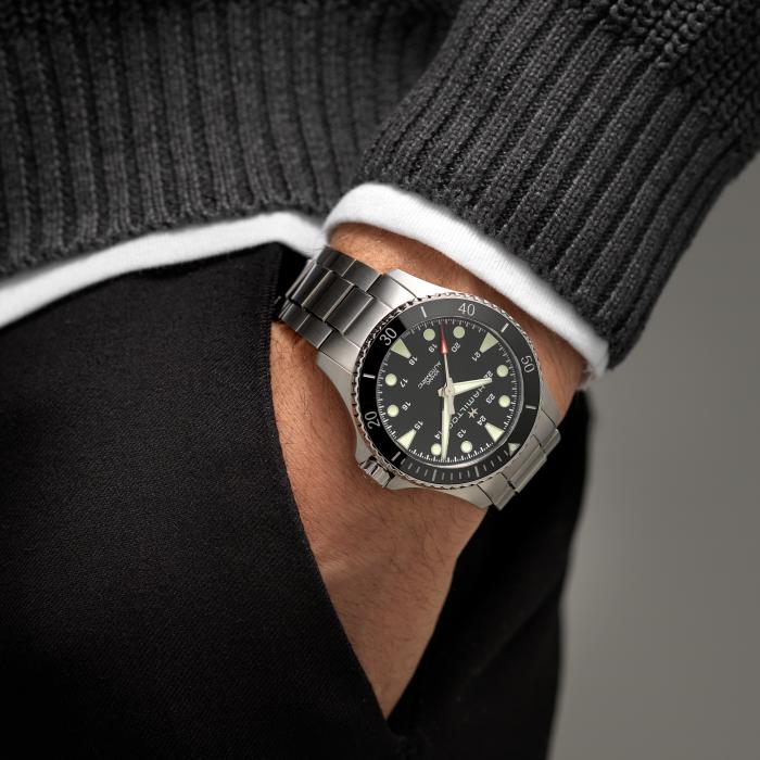 A close-up of a person wearing the Hamilton Khaki Navy Scuba Automatic Watch, featuring a stainless steel band and a black dial. The person's hand is placed in their pants pocket, dressed in a dark sweater with white trim. The watch display includes multiple markings and buttons, highlighting its suitability for deep-sea exploration due to its water resistance.