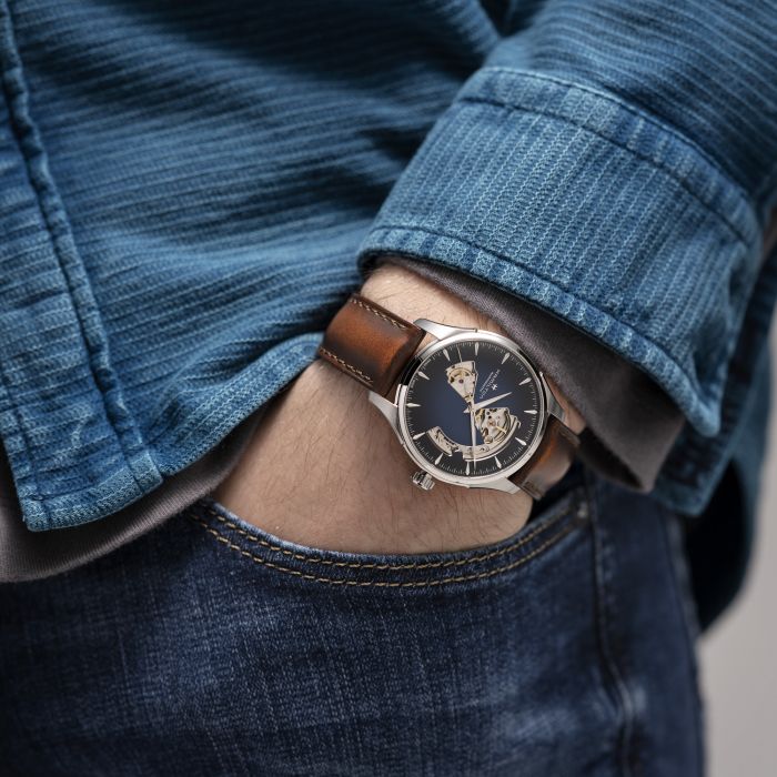 Close-up of a person in a blue long-sleeve shirt and jeans, hand in their right pocket, showcasing the Hamilton Jazzmaster Open Heart Automatic Watch. The wristwatch features a smoked dial with silver-toned hardware and an exposed mechanical movement, complemented by a brown leather strap.