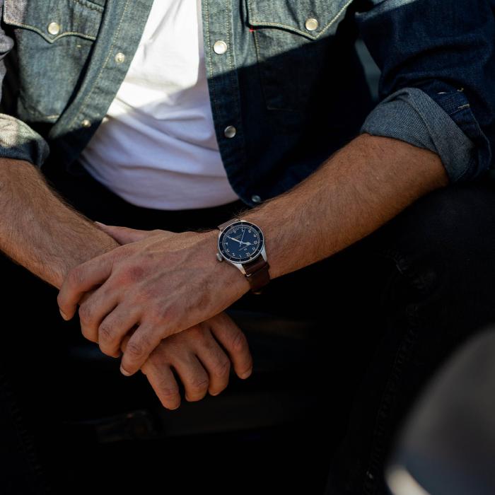 A person is sitting with their hands clasped in front of them, wearing a blue denim shirt over a white T-shirt. On their left wrist, they sport the Hamilton Aviation Pilot Pioneer Automatic Watch (38mm), reflecting Hamilton's aviation heritage. The background is dimly lit.