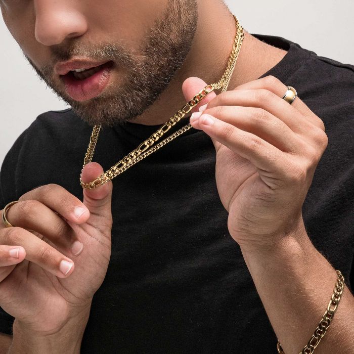 A person with a trimmed beard, wearing a black shirt, showcases an INOX 8mm 18K gold plated Figaro chain necklace by holding it with both hands. The individual also sports gold rings on their fingers against a neutral background.
