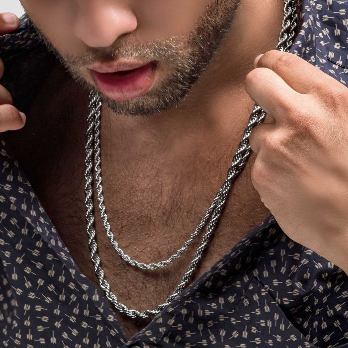 A close-up image shows a person wearing a dark patterned shirt and two men's stainless steel necklaces, one of which is the INOX 4mm Steel Rope Chain | 20". The individual is tugging their shirt open slightly with one hand to prominently display the necklaces. The partial view of the person's face reveals a trimmed beard and slightly open lips.