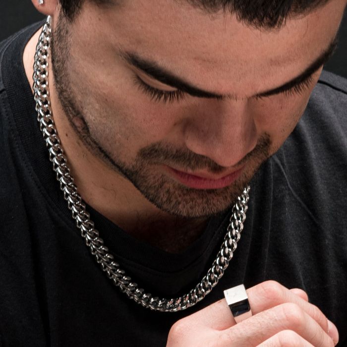 A close-up image features a person with a short beard, wearing an INOX 6mm Steel Franco Chain (22") necklace and holding a ring in their hand. They appear to be looking down at the ring while dressed in a black shirt, with the stainless steel gleam of the men's INOX necklace catching the light.