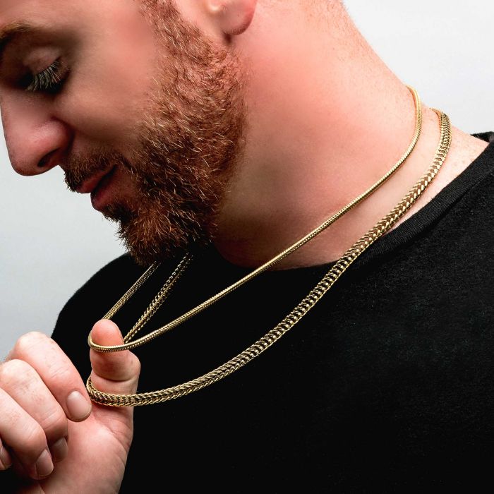 A person with a trimmed beard is shown against a plain background, wearing a black shirt and holding two 3mm 18K Gold Plated Rattail Chains from INOX around their neck. The person looks downward while touching the chains with one hand.