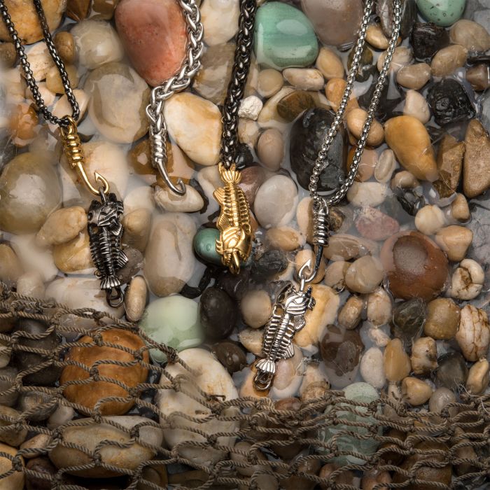 Four fishing hook-shaped pendants, one gold and three silver, rest on a collection of wet, multicolored pebbles with some metal chain links extending above them. An INOX Polished Steel Fishbone Pendant with Hook & Box Chain adds rugged charm with its 24-inch chain. A fishing net is partially visible in the lower part of the image.